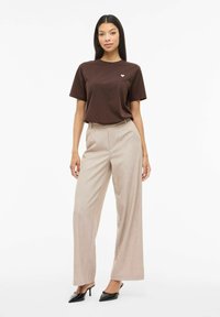 Maglietta di cotone marrone con piccolo logo a forma di cuore, abbinata a pantaloni a gamba larga color beige chiaro. Il look è completato da mule nere con tacco.