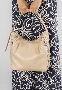Beige leren handtas met laser-gecutte bloempatronen, een gebogen vorm en een enkele schouderband, met een rits en kwastdetail.