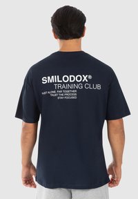 Camiseta de algodón azul marino de manga corta, con un texto en blanco destacado en la parte posterior: "SMILODOX TRAINING CLUB" y frases motivacionales.