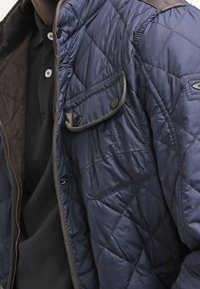 Vatteret mørkeblå jakke med brune detaljer, der har en lomme med knapklap og kontrastsømme på kraven og manchetterne.