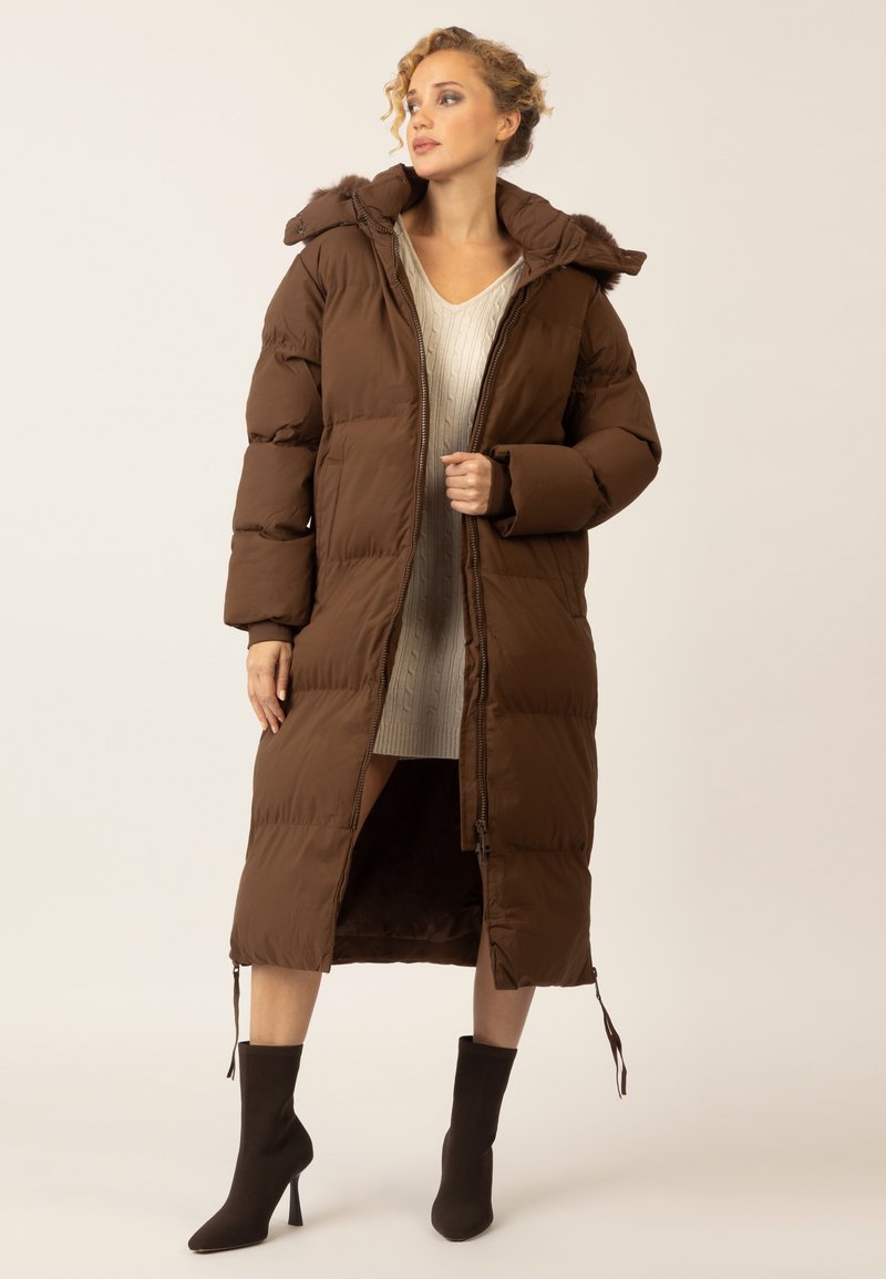 Frau in einem langen braunen Puffercoat über einem cremefarbenen Strickkleid und dunklen, absatzbewehrten Stiefeletten, steht vor einem schlichten, hellen Hintergrund.