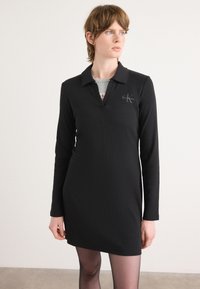 Vestido preto de mangas compridas com um colarinho pontudo e decote em V. Apresenta um pequeno logotipo no peito e uma silhueta ajustada.
