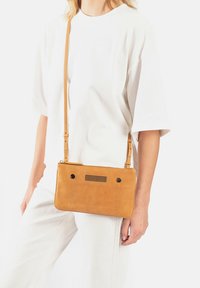 Tan leren crossbody tas met een rechthoekige vorm, ritssluiting, metalen accenten en twee gaten voor het bevestigen van de verstelbare schouderband.