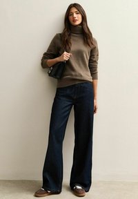Jersey de cuello alto marrón combinado con unos jeans de pierna ancha azul oscuro. El modelo sostiene un pequeño bolso negro y lleva zapatillas slip-on marrones.