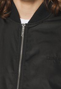 Blouson aviateur noir avec col côtelé, fermeture éclair argentée et détail brodé sur la poitrine. Tissu lisse, design minimaliste.