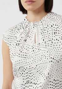 White blouse featuring a gathered neckline, short sleeves, and a keyhole detail. The fabric displays a black abstract pattern of dots and lines.