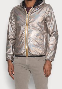 Man in een metallic zilveren hoodie-jack met een rits aan de voorkant en beige broek, staande tegen een effen achtergrond.