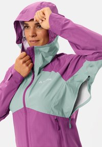 Veste légère avec une partie supérieure violette et une partie inférieure vert menthe ; elle possède une capuche et des poches zippées. Matériau doux et résistant à l'eau.