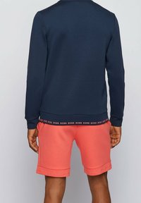 Uomo con pantaloni corti color corallo e felpa blu navy con il testo "BOSS" ripetuto sulla banda e sui polsini dei pantaloni, in piedi con la schiena rivolta verso l'osservatore.