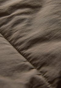 Tissu marron avec une surface texturée, présentant des ondulations subtiles et une couture matelassée le long d'un bord. Matériau doux et léger.