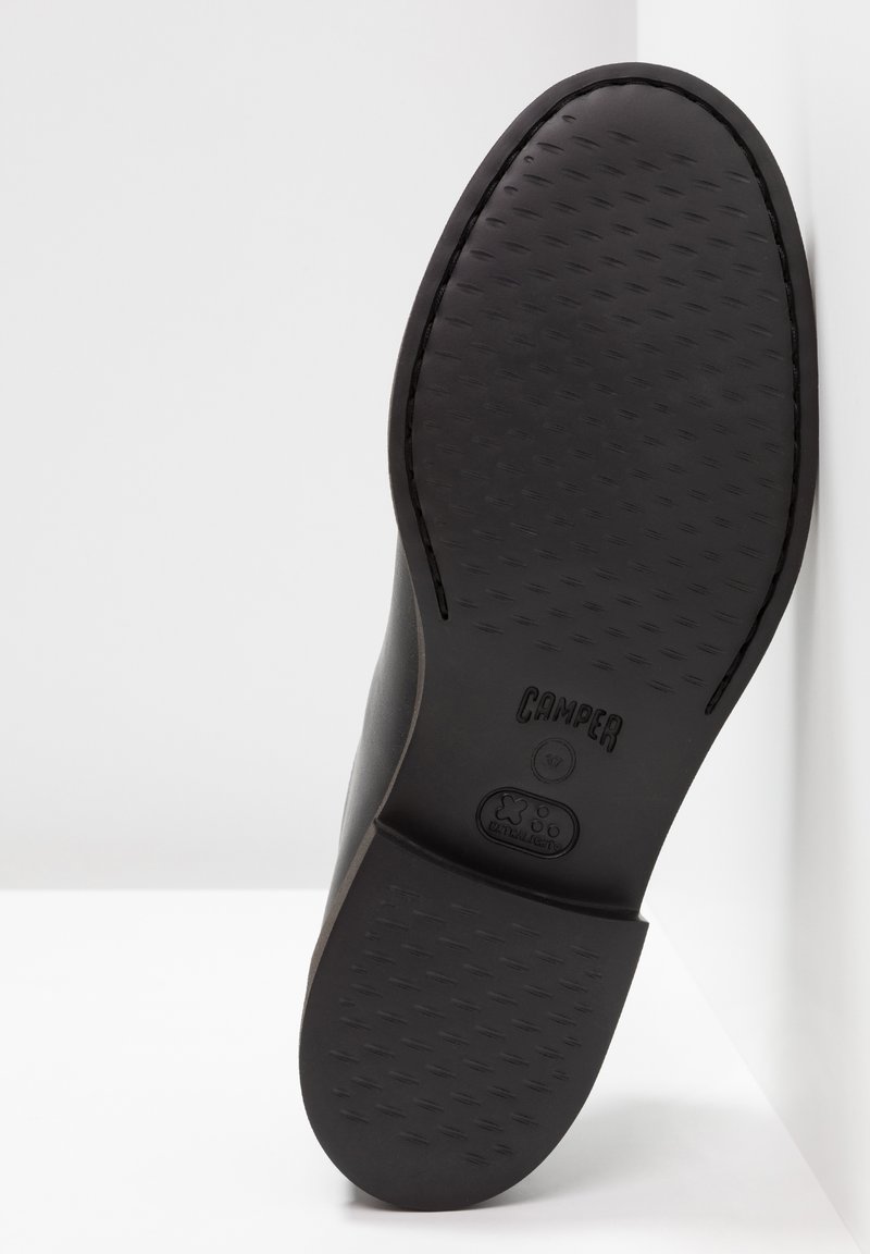 Schwarze Schuhsohle mit strukturiertem Muster und abgerundeten Kanten. Mit eingraviertem Markennamen "Camper" und einem Logo in der Nähe der Ferse.