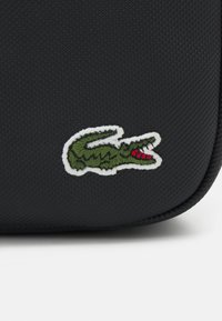 Borsa in materiale nero testurizzato con un logo di coccodrillo ricamato verde e rosso su sfondo bianco. Design compatto con bordi arrotondati.
