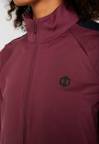 Maronfarbener Zip-up-Jacke aus glattem, leichtem Material. Verfügt über einen hohen Kragen und schwarze Akzente auf den Schultern, mit einem sechseckigen Logo auf der Brust.