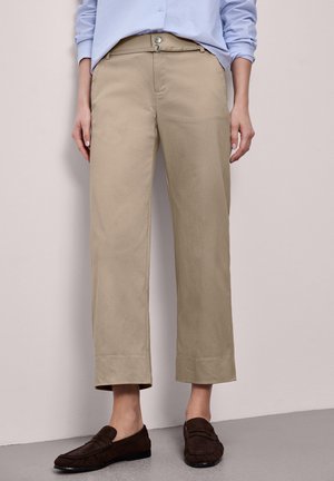 Kvinde iført beige bukser med lige ben, lyseblå skjorte med knapper og oprullede ærmer samt mørkebrune loafers, stående op ad en ensfarvet væg.