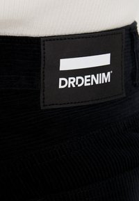 Tissu en velours côtelé noir avec une texture côtelée ; comporte une étiquette rectangulaire avec le texte blanc "DRDENIM" et une ligne blanche horizontale.