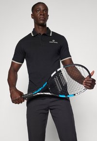 Melnas polo krekls ar baltu un pelēku svītrotu apkakli, ar Sergio Tacchini logotipu. Tur rokās melnu tenisa raketi ar zilām akcentiem.