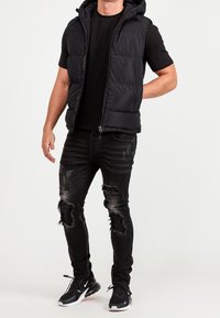 Uomo che indossa un giubbotto smanicato imbottito nero, una t-shirt nera, jeans neri strappati con toppe a forma di stella e sneakers Nike nere, in piedi davanti a uno sfondo bianco.