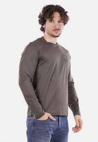 Koszula z długim rękawem w ciemnoszarym kolorze z bawełny, z okrągłym dekoltem i kieszonką na piersi z małym akcentującym wzorem. Noszona z niebieskimi dżinsami.