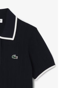 Polo bleu marine avec des bords blancs et une texture côtelée. Présente un petit logo crocodile vert brodé sur la poitrine.