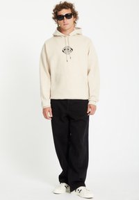 Beige hoodie met zwart grafisch ooglogo, voorzak en trekkoord. Gecombineerd met zwarte broek en witte sneakers met zwarte accenten.