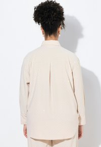 Camisa de manga larga beige con sutiles lunares, cuello con botones y pliegue en la parte posterior central. Tejido suave con un corte holgado.