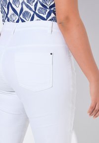 Jeans de mezclilla blanca con un diseño ajustado, que cuentan con un único bolsillo trasero con un remache negro, combinados con una blusa de estampado azul marino.