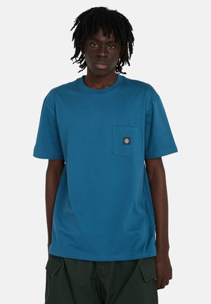 Junge Person mit dunkler Haut und Dreadlocks, die ein blaues T-Shirt mit Taschen und dunkelgrüne Cargo-Hosen trägt und vor einem schlichten Hintergrund steht.