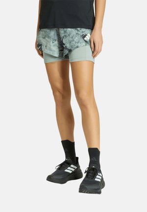 Piernas con pantalones cortos deportivos grises estampados sobre pantalones cortos de compresión gris claro más largos, con calcetines negros hasta la mitad de la pantorrilla y zapatillas negras para correr.