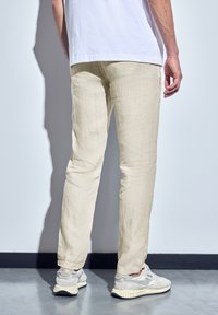Pantalons en lin beige clair, coupe droite, tissu texturé. Portés avec des chaussures claires, debout devant un mur neutre.