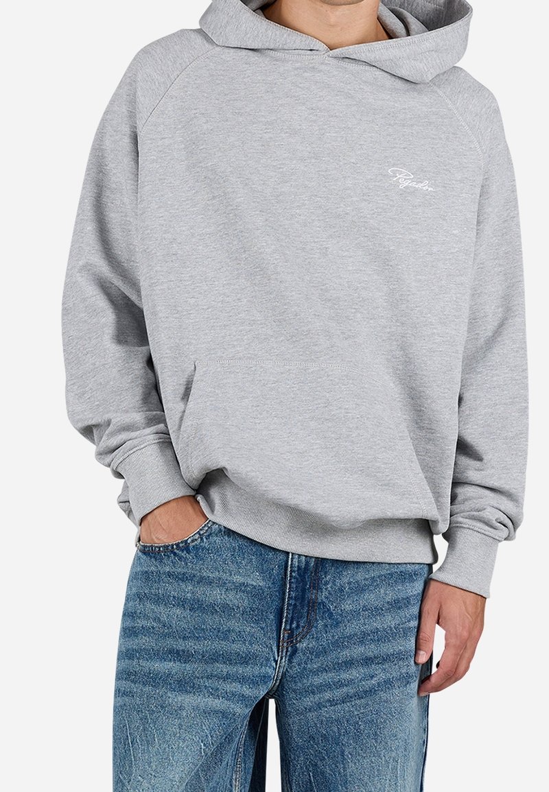 Homme portant un sweat à capuche gris clair avec poche kangourou et un jean bleu, main dans la poche, sur fond uni.
