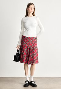 Jupe à carreaux rouges avec des plis, associée à un haut à manches longues blanc. Les accessoires comprennent un petit sac noir et des chaussures noires à lanières.