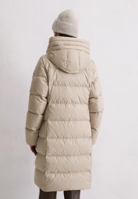 Henkilö, joka on pukeutunut pitkään beigeen tikattuun takkiin, jossa on huppu, sekä yhteensopivaan beigeen neulottuun pipoon, seisoo vaaleaa taustaseinää vasten.