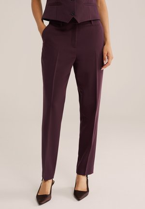 Femme portant un pantalon violet foncé sur mesure avec poches et un gilet assorti, associé à des talons pointus foncés.