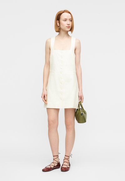 Κοντό φόρεμα σε κρεμ απόχρωση με τετράγωνο ντεκολτέ, κουμπιά μπροστά και εφαρμοστή γραμμή. Η μοντέλα κρατάει μια πράσινη τσάντα και φοράει καφέ παπούτσια με κορδόνια.
