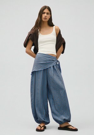 Junge Frau trägt ein weißes Tanktop, eine braune Jacke, blaue karierte weite Hosen, die an der Taille gebunden sind, und schwarze Sandalen und steht ruhig.