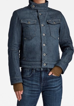 Mørkeblå denimjakke med knaplukning, brystlommer med klap og høj krave båret over brune fingerløse handsker og matchende denimjeans.