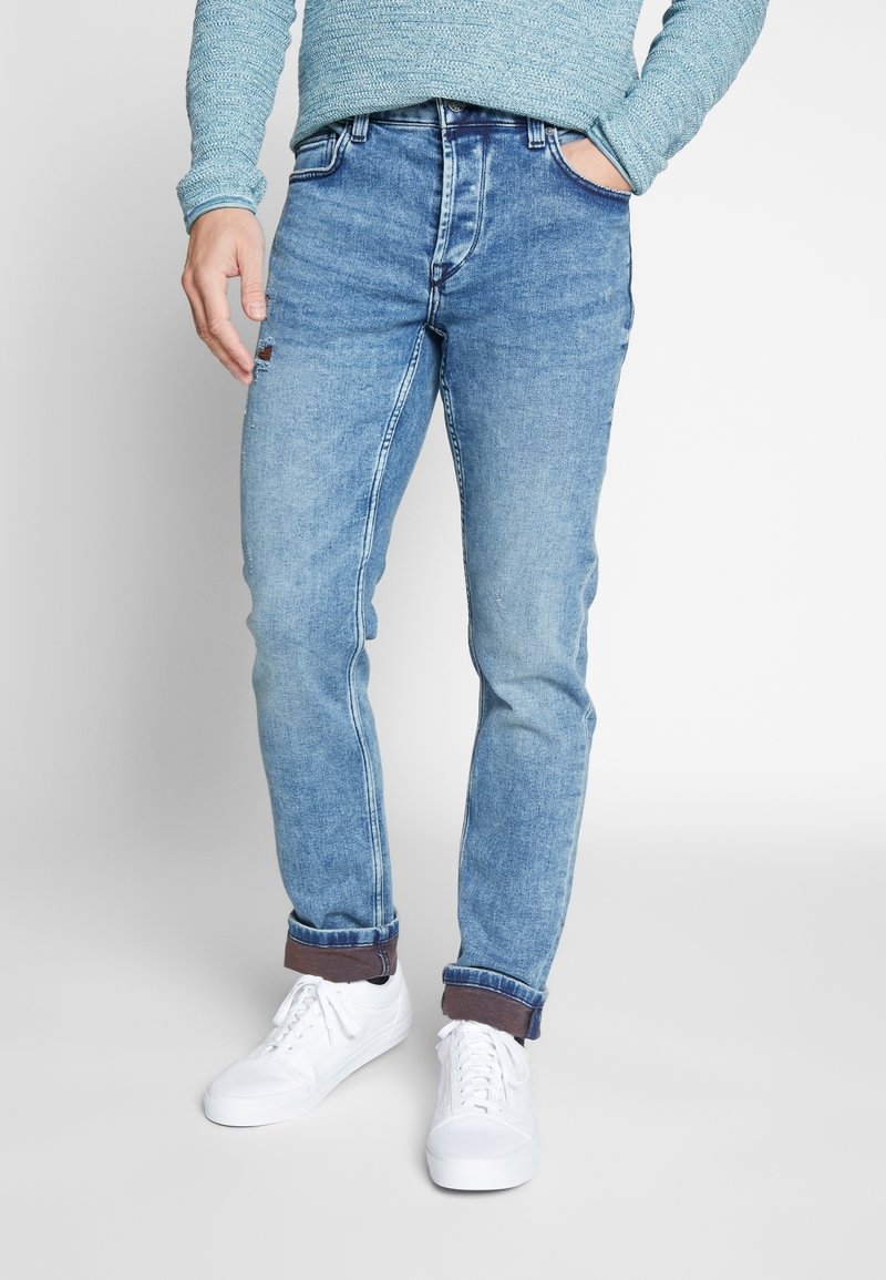 Jeans de mezclilla azul claro con un corte ajustado, que presentan un diseño clásico de cinco bolsillos, puños enrollados y sutiles detalles de desgaste. Llevados con zapatillas blancas.