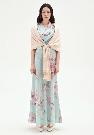 Femme aux longs cheveux foncés portant une robe maxi fleurie bleu clair et un châle beige noué à l'avant, debout sur un fond blanc.