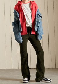 Persona vistiendo una sudadera roja con cremallera sobre una camiseta blanca y una chaqueta de mezclilla, pantalones negros y zapatillas en blanco y negro, de pie en el interior.