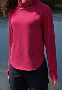 Maglia a maniche lunghe in tessuto rosa vivace con scollo a cowled, caratterizzata da una texture morbida e un orlo arrotondato. Abbinata a pantaloni scuri.