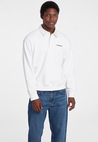 Fehér pulóver, póló gallérral, fekete "Guess Jeans" logóval. Kék farmernadrággal viselve, laza szabással.