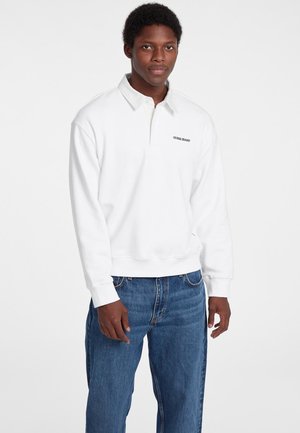 White sweatshirt with a polo collar, featuring a black "Guess Jeans" logo. Worn with blue denim jeans, showing a relaxed fit.