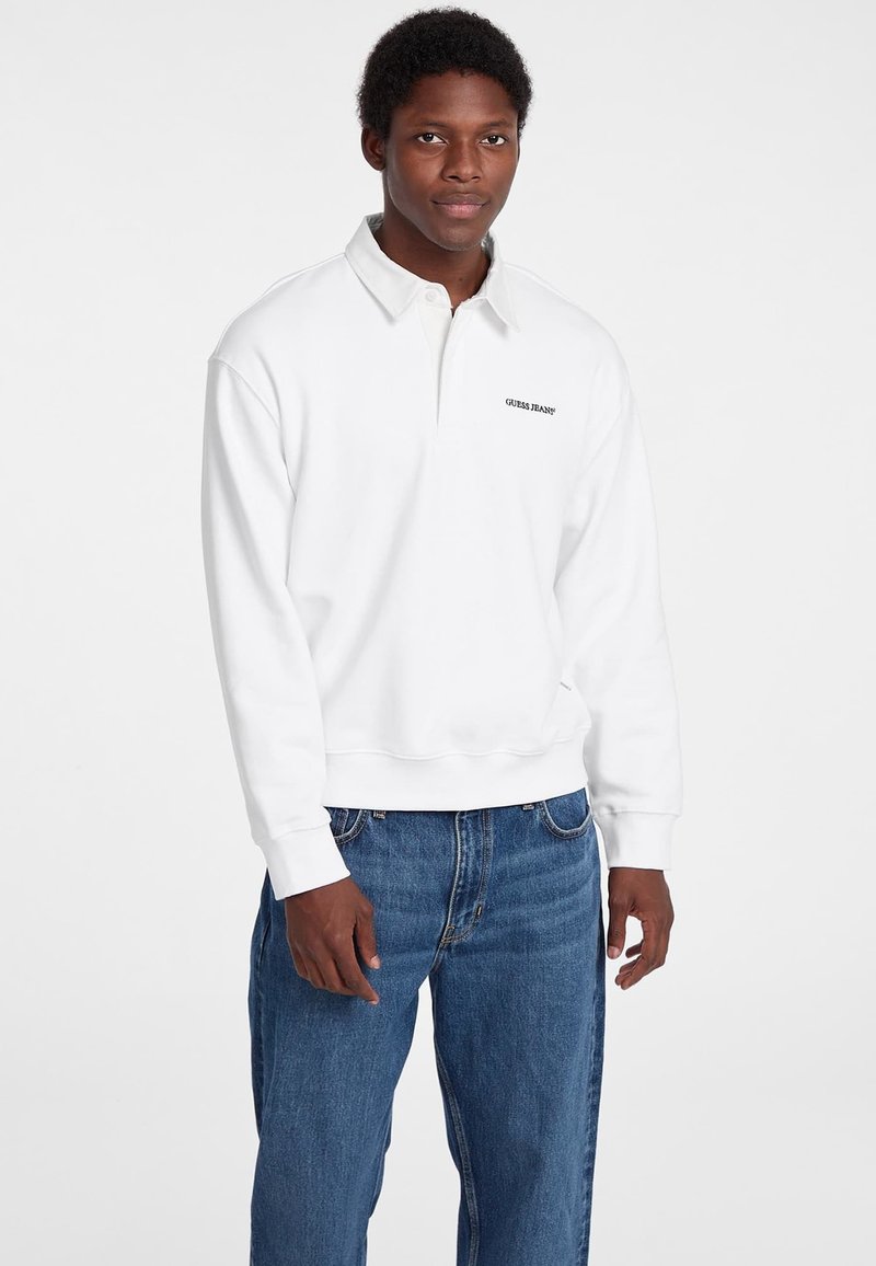 Fehér pulóver, póló gallérral, fekete "Guess Jeans" logóval. Kék farmernadrággal viselve, laza szabással.