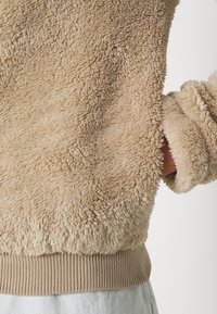 Person trägt eine beige, flauschige Fleecejacke mit geripptem Saum und Ärmelbündchen, die Hand in die Seitentasche gesteckt, vor einfachem hellem Hintergrund.