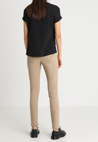 Kvinna klädd i en svart kortärmad skjorta, beige slim-fit byxor och svarta snörskor, som står mot en enkel ljus bakgrund.