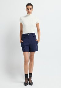 Vrouw die staat met handen in haar zakken, gekleed in een witte gebreide top met korte mouwen, marineblauwe high-waisted shorts, doorzichtige zwarte sokken en zwarte hakken.