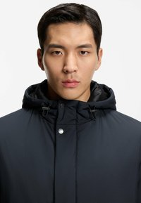 Chaqueta acolchada negra con capucha, cierre de botones y cordones ajustables. Textura suave y diseño minimalista.