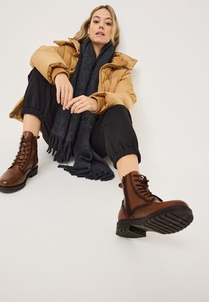 Braune Leder-Schnürstiefel mit strukturierter Oberfläche, kombiniert mit schwarzen schmalen Hosen und einem dunkelgrauen Schal, vor einem neutralen Hintergrund.