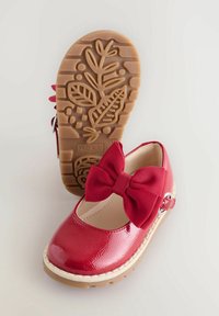 Scarpa in pelle lucida rossa con un grande fiocco testurizzato, punta arrotondata e suola in gomma beige con motivo a foglia. Cinghia regolabile con fibbia.