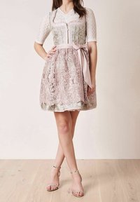 Vrouw in een roze kanten jurk tot op de knie met een bloemen-corsage en een gestrikte sjerp, gecombineerd met zilveren enkelbandhakken.